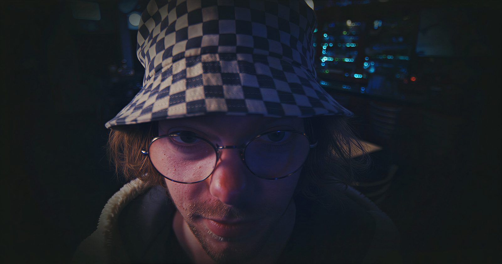 A man in a checked bucket hat and glasses, lit blue, looking off to one side.