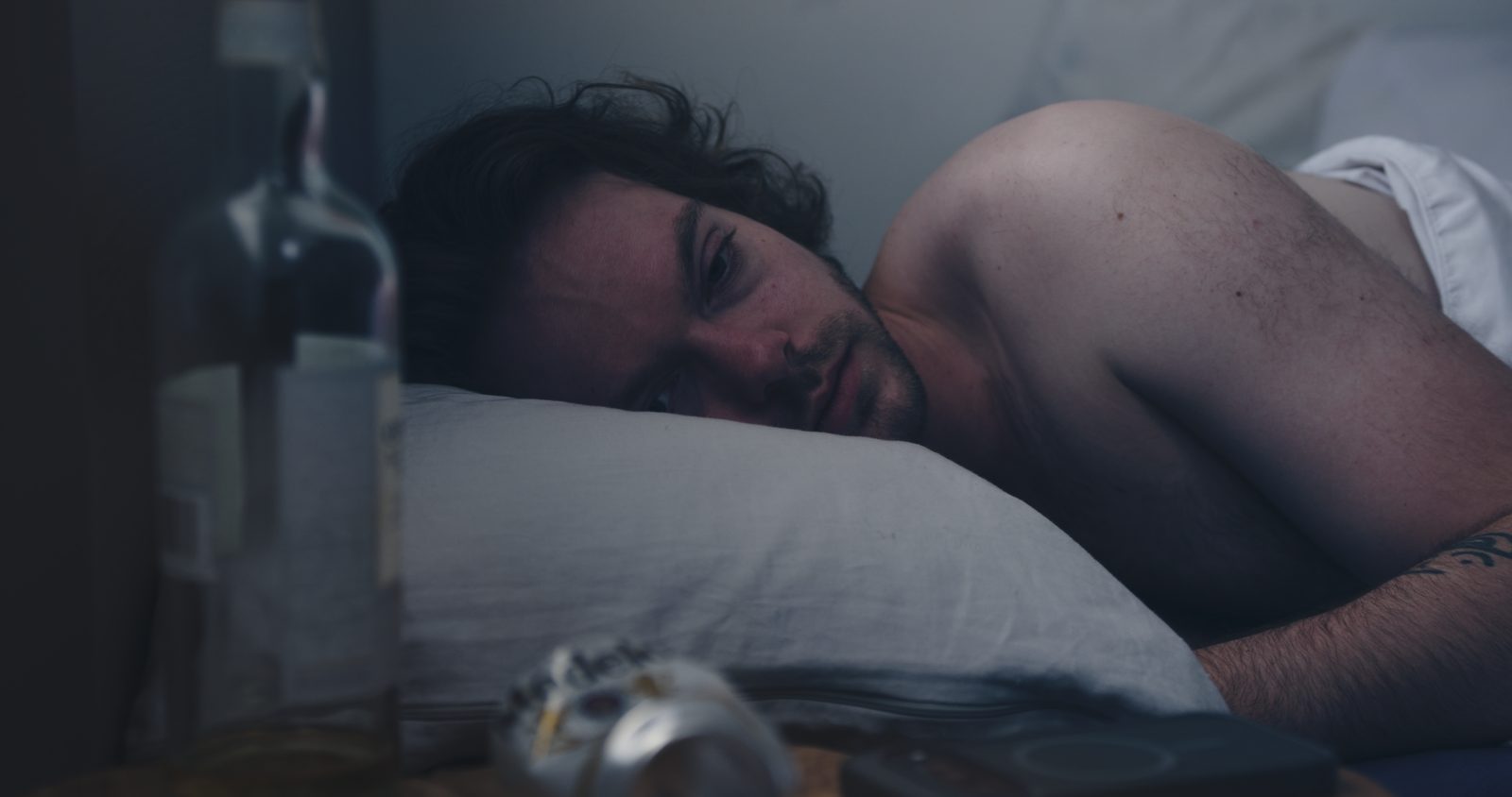 A man lying awake in bed, a bottle in the foreground.