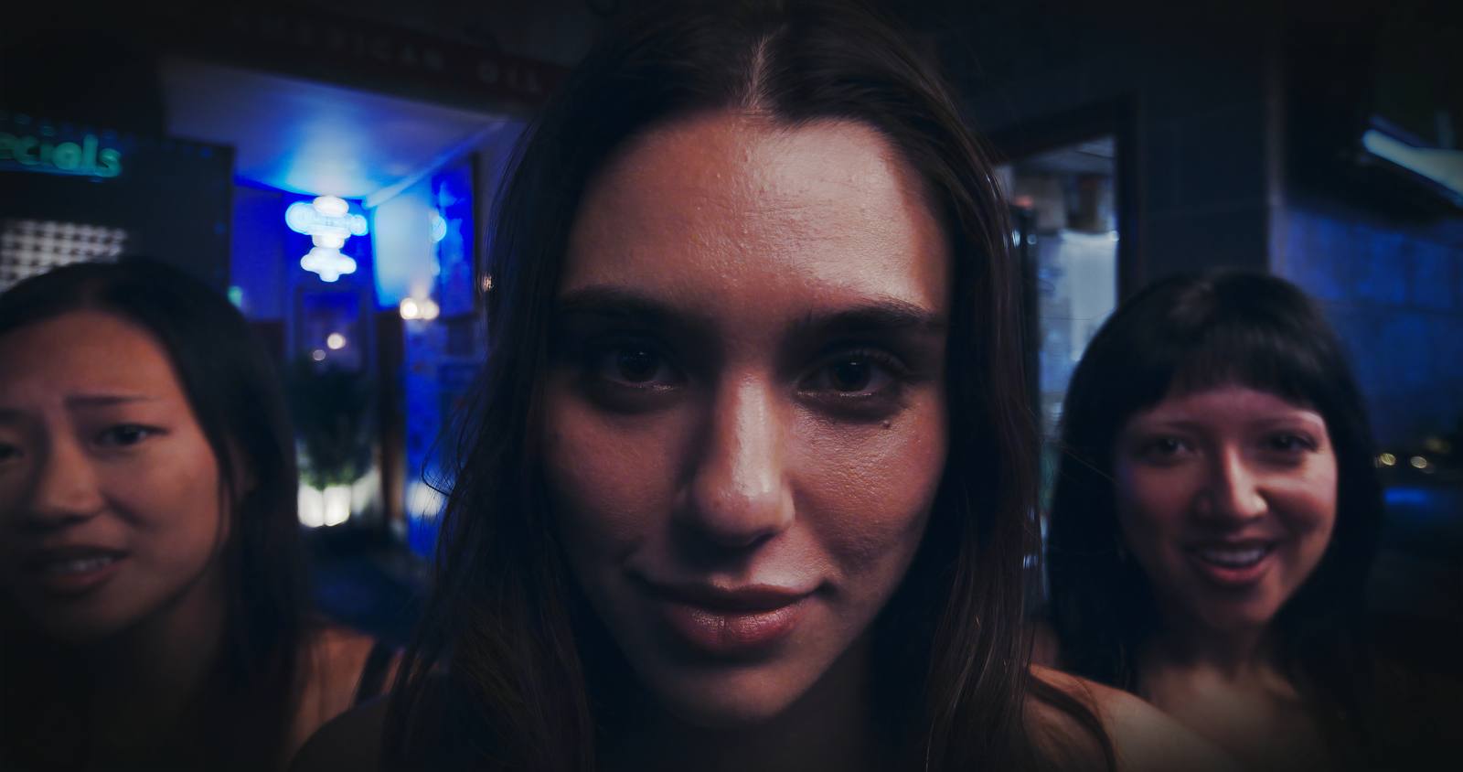 Three young women in a bar lit blue, the nearest looking straight at camera.