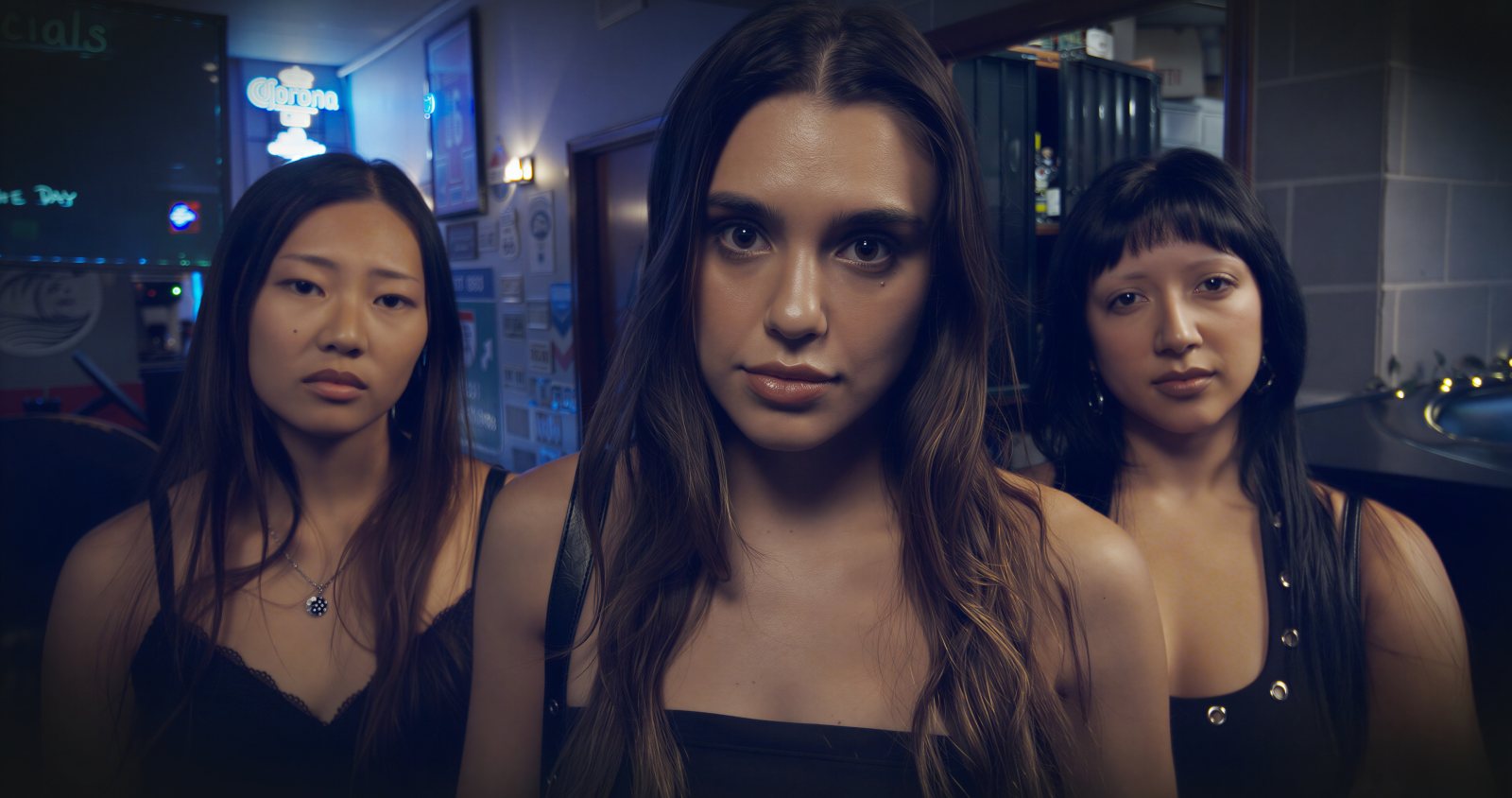 Three young women standing side by side in a bar lit blue, all three looking straight at camera.