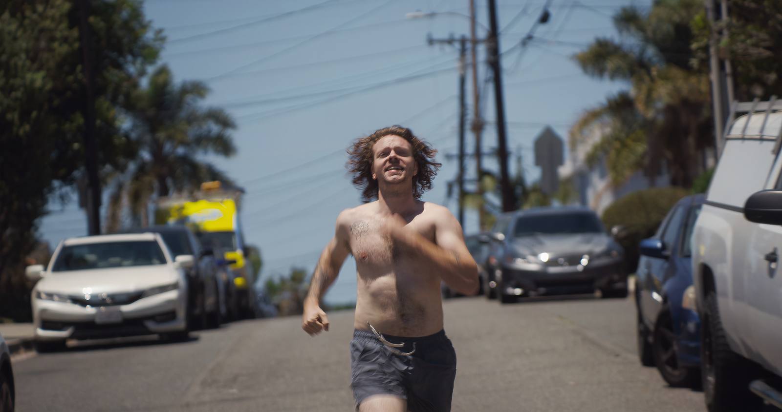 A shirtless man running down the middle of a suburban street in bright daylight, parked cars on both sides.
